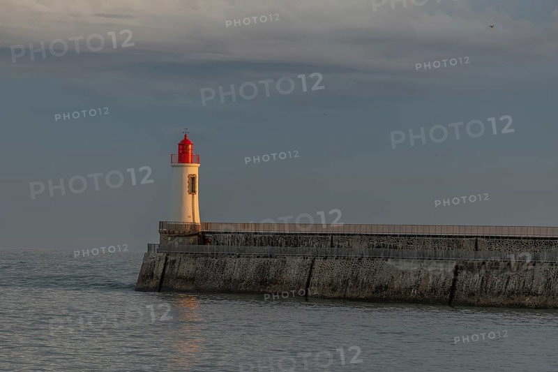 A lighthouse stands on a pier and casts its light over calm waters at dusk. The sky is filled with dramatic clouds, adding to the tranquil atmosphere of the coastal landscape. Les Sables d'Olonne, Vendee, France
