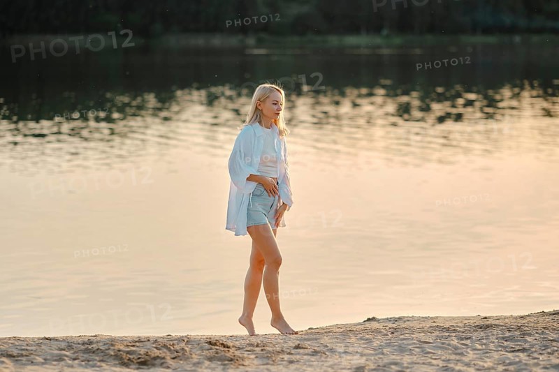 A woman strolls barefoot on a sandy beach by the lake, enjoying the serene atmosphere as the golden hour casts a warm glow over the water and landscape. The soft light enhances the peaceful scenery