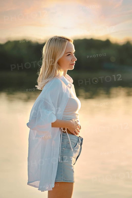 A young woman stands by a calm lake during sunset, feeling the gentle breeze. Her casual outfit reflects the relaxed atmosphere, with the sun casting warm hues on the water and surroundings