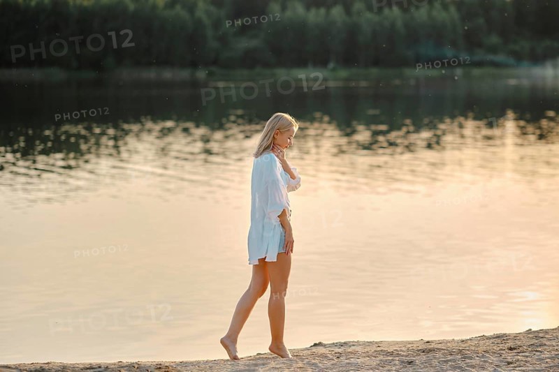 Pensive woman strolls barefoot on the sandy beach beside a serene lake. Golden sunlight reflects off the water as trees stand in the background, creating a calm and soothing atmosphere during sunset