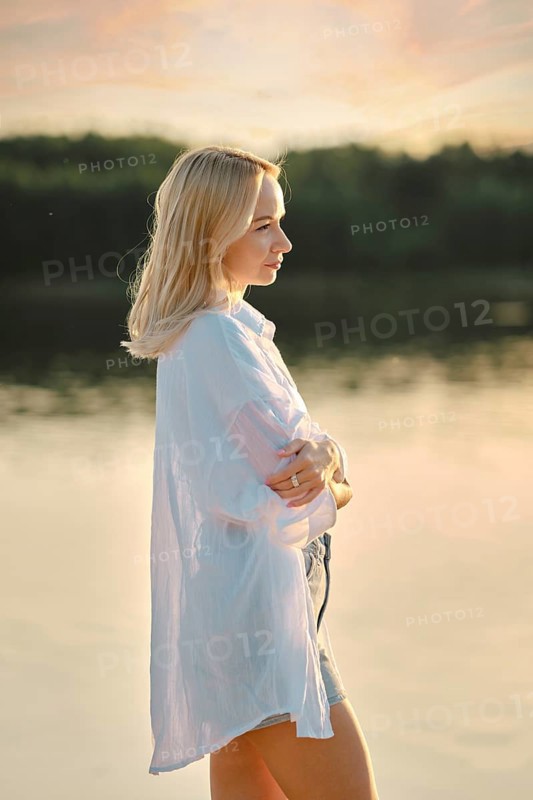 A young woman stands pensively by a calm lake at sunset, dressed in a light shirt and shorts. The warm glow of the setting sun reflects on the water, creating a peaceful atmosphere