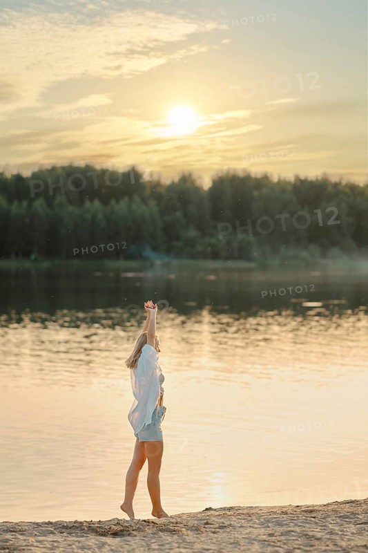 A woman stands on the sandy shore of a tranquil lake, raising her hand joyfully towards the setting sun. The golden light reflects off the water, surrounded by lush greenery
