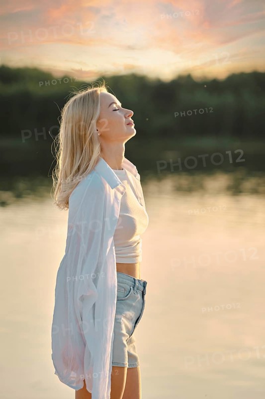 A woman stands by the calm lake, feeling the gentle breeze as she admires the colorful sunset sky. She wears a light shirt and shorts, embracing the peaceful atmosphere of the evening