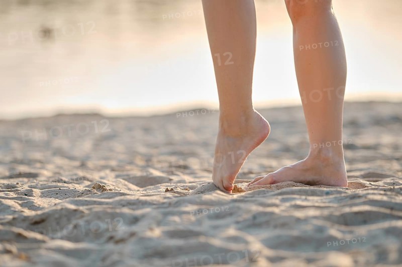 A person stands on soft, golden sand with bare feet, enjoying the warm texture as the sun sets in the background. The peaceful beach atmosphere enhances the sense of relaxation and freedom
