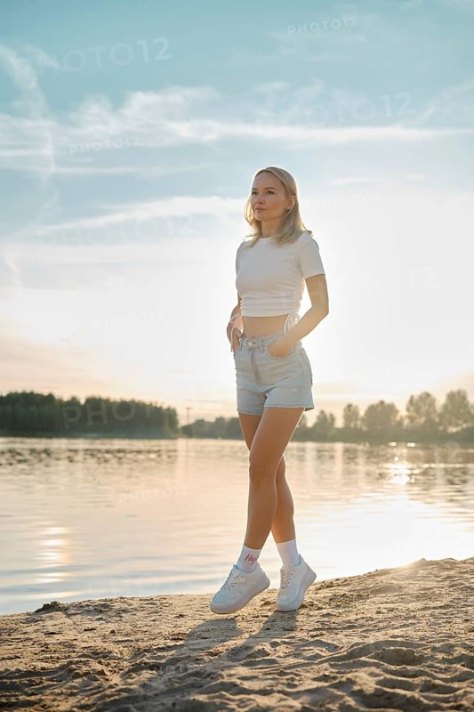 A young woman walks on tiptoes on a sandy shore as the sun sets over the lake. Her casual outfit complements the serene atmosphere, with soft light illuminating the scene