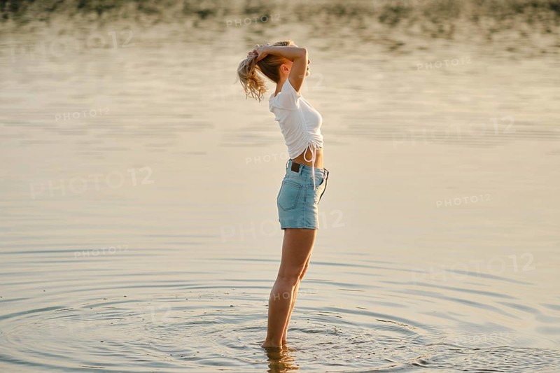 A woman stands in shallow water in profile, basking in the warm glow of sunset. She raises her hands and gathers her hair in a ponytail