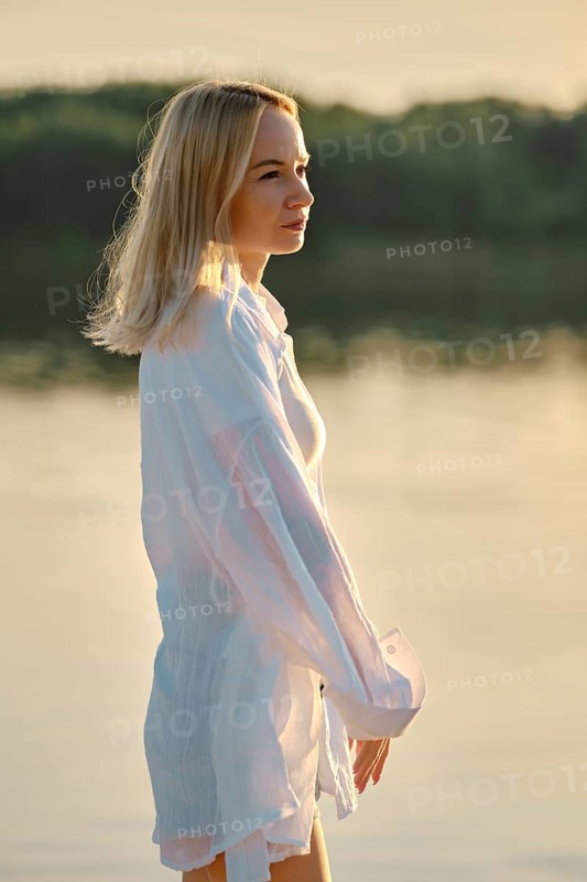Woman with light blonde hair gazes thoughtfully into the distance while standing near tranquil water. The sun sets behind trees, creating a warm, golden light that enhances the peaceful atmosphere