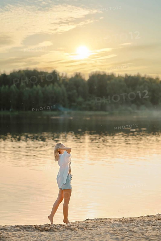 A barefoot woman walks along the sandy shore of the forest lake. She set her face to the warm rays of the sunset sun