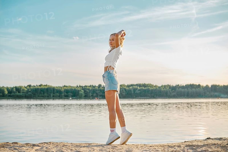 A young woman walking along a tranquil lake at sunset, dressed in casual summer attire. She stretches her arms blissfully, surrounded by lush greenery and calm water reflecting the sky