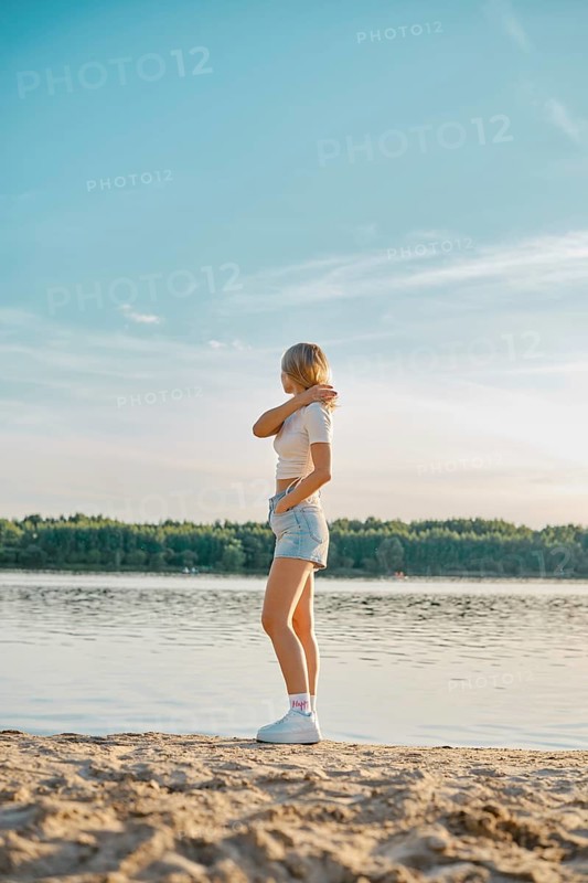 A young woman stands on the sandy shore of a tranquil lake, gazing at the water while wearing casual summer attire. The sun sets in the background, painting the sky with warm colors