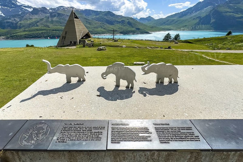 View from viewing platform to Mont-Cenis Pyramid Museum with stylised pyramid Museum in memory of Egypt campaigns of Emperor Napoleon Bonaparte and time in front of construction of reservoir, behind it Lac du Mont Cenis, in front on parapet small stylised figures of elephants Symbols for crossing of Alps Crossing of Alps by Hannibal, Mont Cenis Massif, Massif du Mont-Cenis, Graian Alps, Lanslebourg, Rhone-Alpes, France