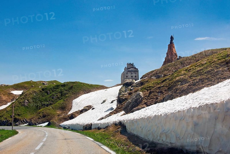 View from last bend of alpine road pass road mountain road in front of pass summit of Little Saint Bernard Pass Colle del Piccolo San Bernardo, Col du Petit Saint Bernard Little Saint Bernard Pass in Graian Alps, top right statue of monk Saint Bernard of Menthon on large pedestal made of natural stones, top centre historic renovated reopened hotel Hospice l'Hospice de Petit Saint Bernard, Département Savoie, France