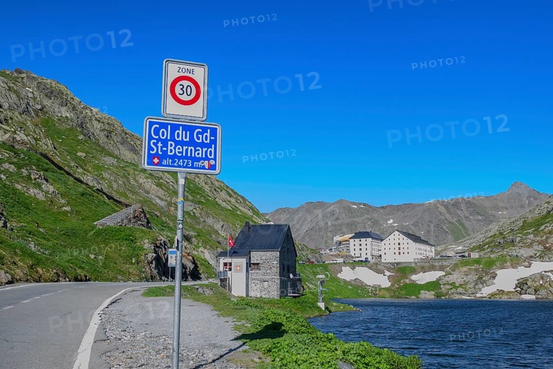 Traffic sign Speed limit sign 30 km/h below information Col du Gd-St.Bernard Grand Saint Bernard and altitude 2473 metres, behind it former border building at border crossing from Italy to Switzerland, in the background historic Auberge Herberge Hospice on top of the pass of Grand Saint Bernard, Saint-Rhémy-en-Bosses, Aosta Valley region, Italy