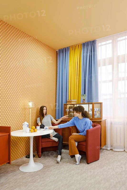 Two women are seated in a bright room, engaged in a lively discussion. One woman reaches out with a playful expression while the other listens attentively