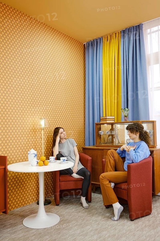 Two friends are sitting comfortably in a living room with bold wallpaper and bright curtains. They engage in a conversation, discussing female problems