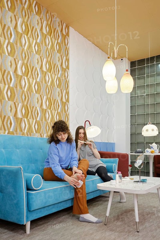 Two friends enjoy a cozy moment in a modern lounge, sitting on a colorful sofa. One woman removes rough skin on the foot, and the other uses a nail file