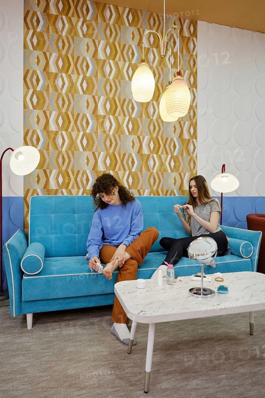 Friends spend a pleasant afternoon in a modern lounge, sitting on a turquoise sofa. They engage in conversation while one polishes her nails and the other removes dead skin of heel