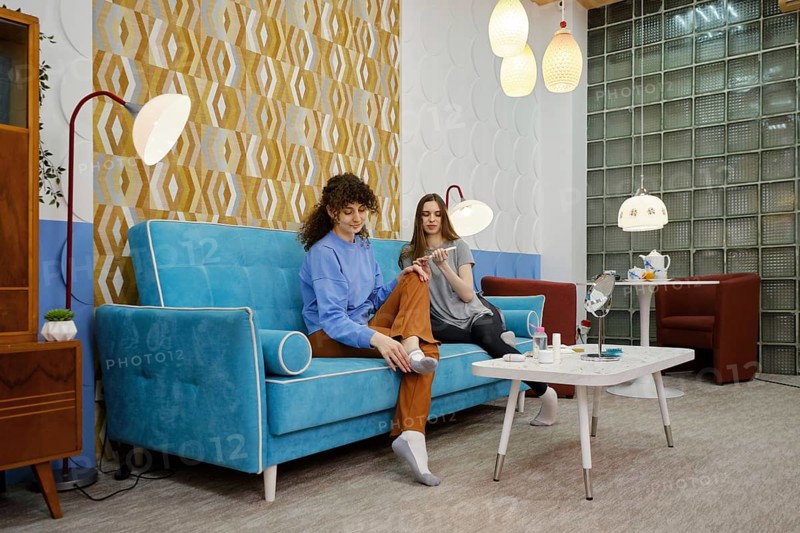 Two women are seated on a blue sofa in a living room, enjoying each other's company while engaging in beauty procedures