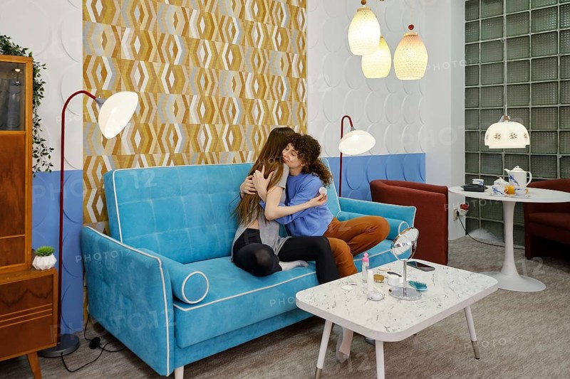 Two friends embrace on a blue couch in a beautifully decorated room featuring warm light fixtures and vibrant wallpaper, conveying a sense of comfort and connection during their time together