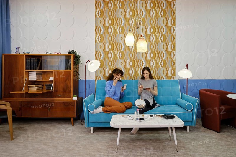 Two friends are seated on a vibrant blue couch in a modern living room, engaged in light conversation while applying make-up