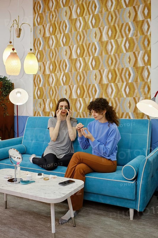 Two friends are enjoying a relaxed moment on a blue couch, engaged in a beauty session. They are applying patches and cream while laughing and sharing tips in a cozy, modern living space