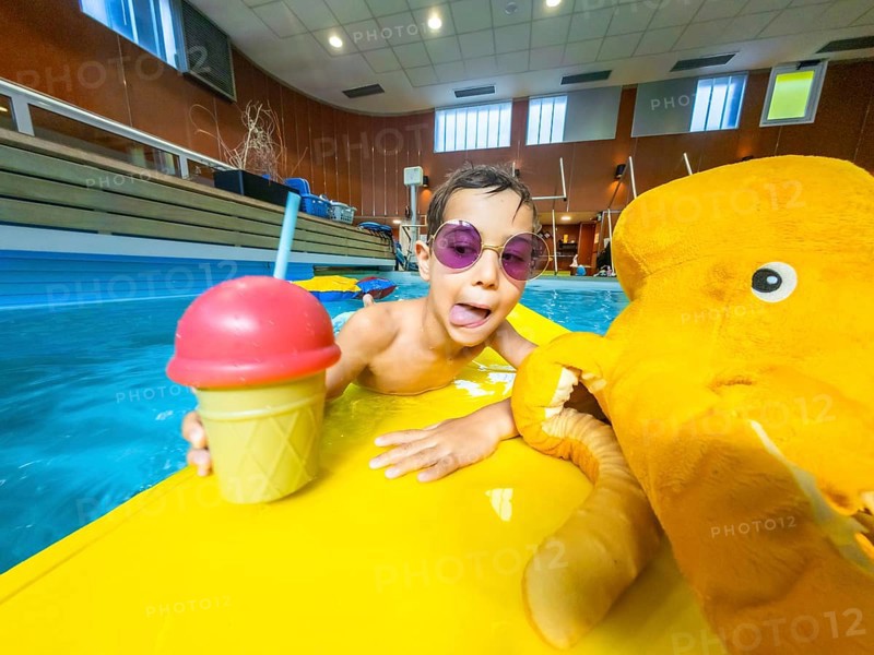 Boy with sunglasses sticking out his tongue, holding a refreshing drink and a plush toy while floating on a yellow inflatable mattress in a pool
