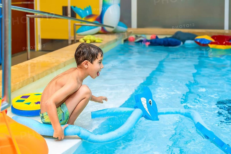 Happy child crouching at the edge of an indoor swimming pool, playing with a pool noodle shaped like a shark
