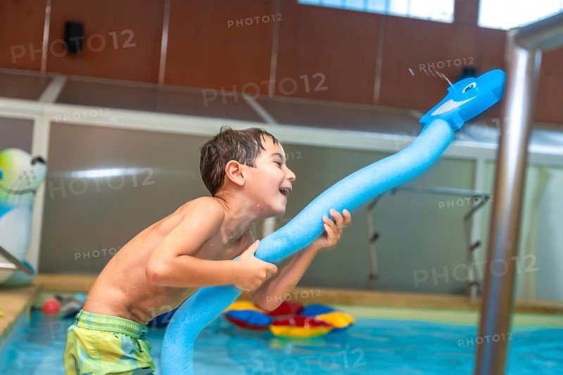 Happy child playing with a pool noodle shaped like a whale and spraying water in an indoor swimming pool