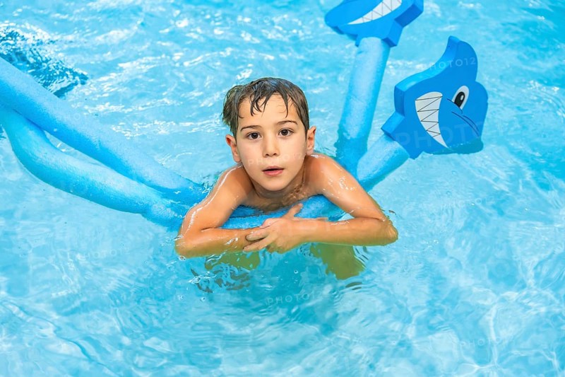 Young boy playing with a shark shaped pool noodle float in a blue swimming pool, enjoying his summer vacation