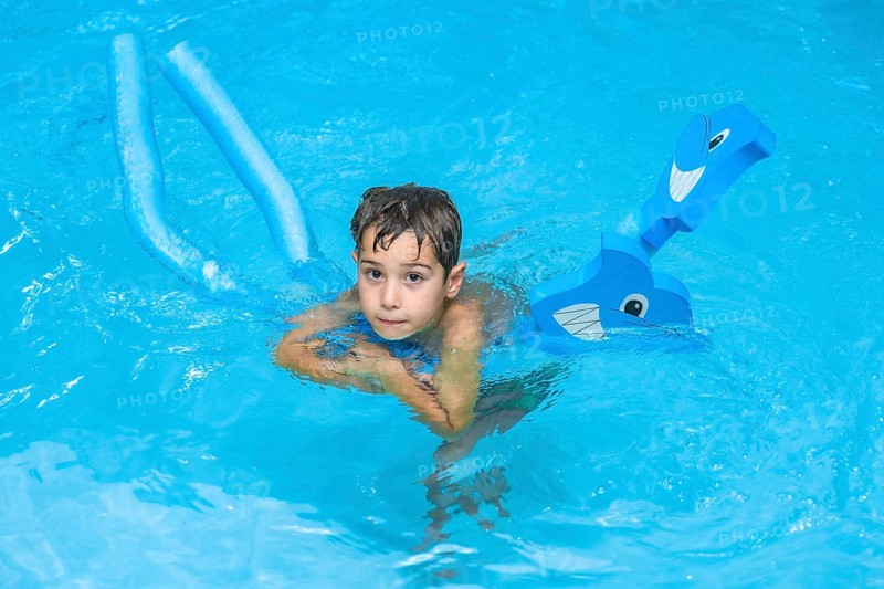 Boy swimming and playing with pool noodles and inflatable toy in a blue swimming pool during summer