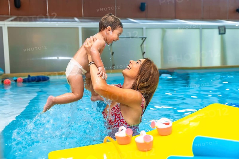 Happy mother lifting her laughing baby out of the water in an indoor swimming pool next to floating toys
