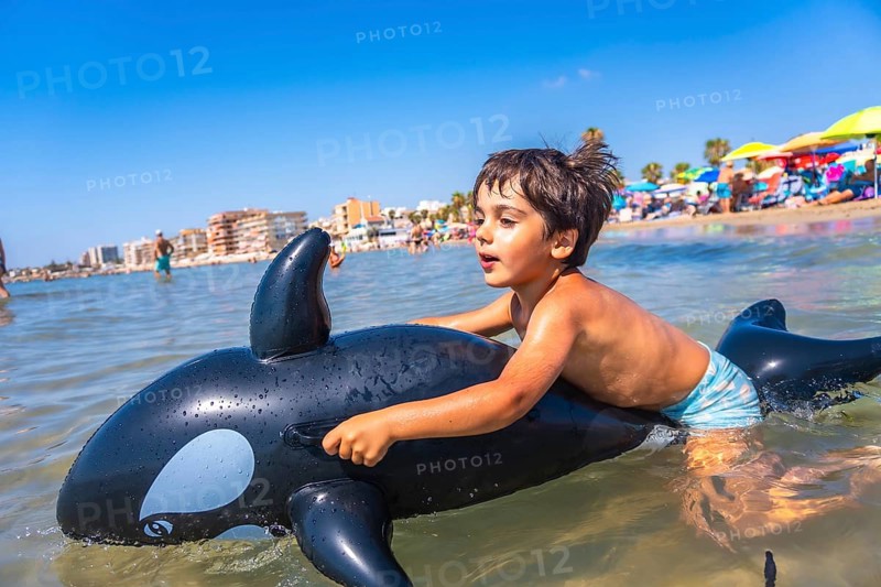Happy child riding on an inflatable orca toy in the sea, splashing through waves and enjoying a joyful summer vacation by the beach