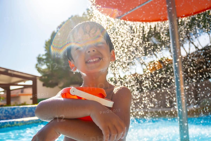 Smiling child holding a bucket and enjoying refreshing shower in a swimming pool on a sunny summer day