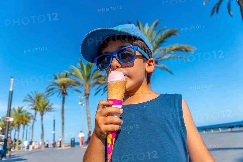Young boy wearing sunglasses and cap enjoying a refreshing ice cream cone on a sunny summer day at the beach, with palm trees in the background
