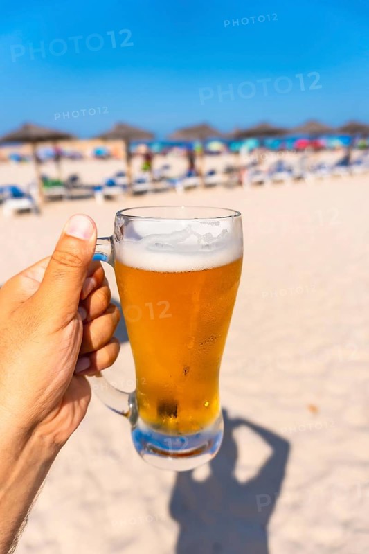 Hand holding glass of cold beer on beach with blurred beach umbrellas and sunbeds in background