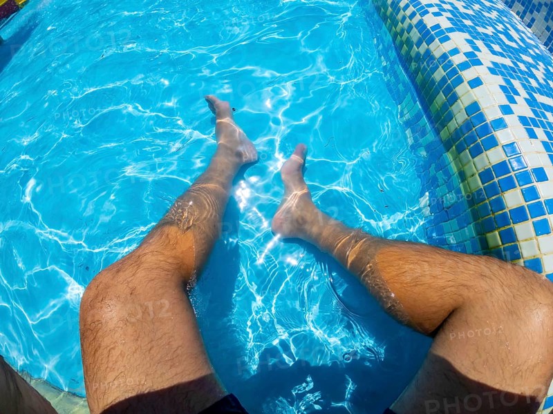 Point of view shot capturing a man relaxing in a swimming pool, legs submerged in the refreshing turquoise water under the sunny sky