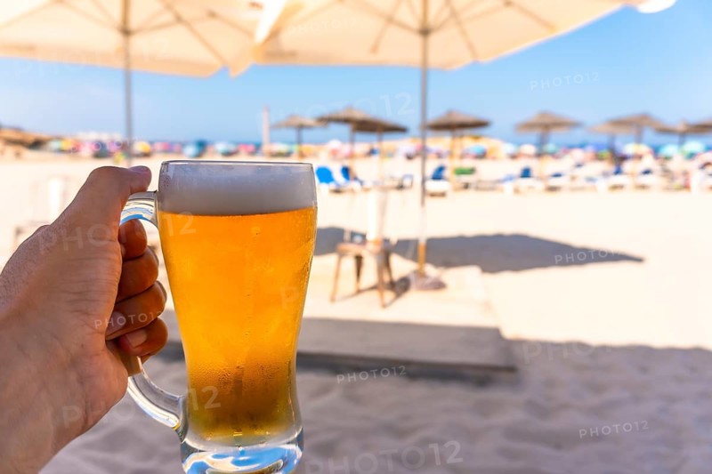 Tourist enjoying refreshing beer on sunny beach with blurred umbrellas and ocean, perfect summer vacation concept