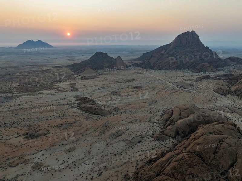The isolated Spitzkoppe mountain (1728 m) - on the right - majestically rises above the surrounding desert plains. In the distance the coast and the Atlantic Ocean. At sunset. Aerial view. Drone shot. Damaraland, Namibia