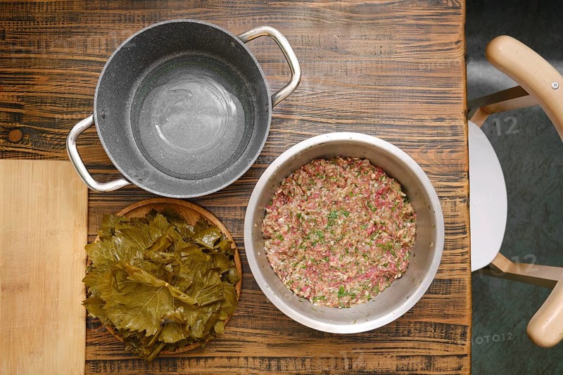 A bowl of prepared ground meat and rice mixture sits next to a plate of grape leaves ready for wrapping on a rustic wooden table