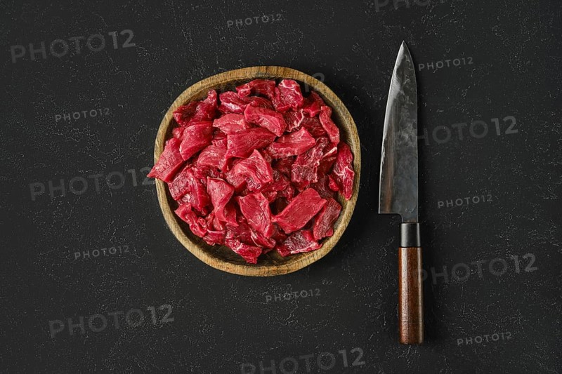 A wooden bowl holds freshly cut pieces of raw beef, showcasing a vibrant red color. Beside the bowl, a sharp knife rests on a dark textured surface, ready for preparation or cooking