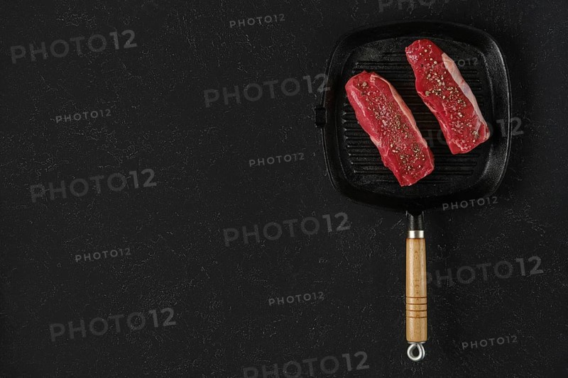 Two raw beef steaks on a preheated griddle pan, seasoned with spices, in a home kitchen setting. The black surface is for text