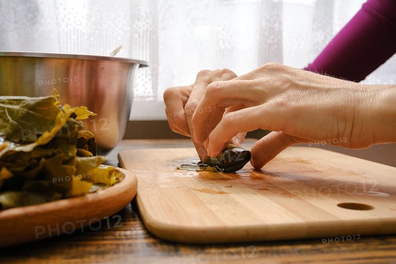 Hands delicately wrap seasoned filling in grape leaves on a wooden cutting board. Fresh ingredients and a mixing bowl are visible, showcasing the traditional culinary process of making dolma