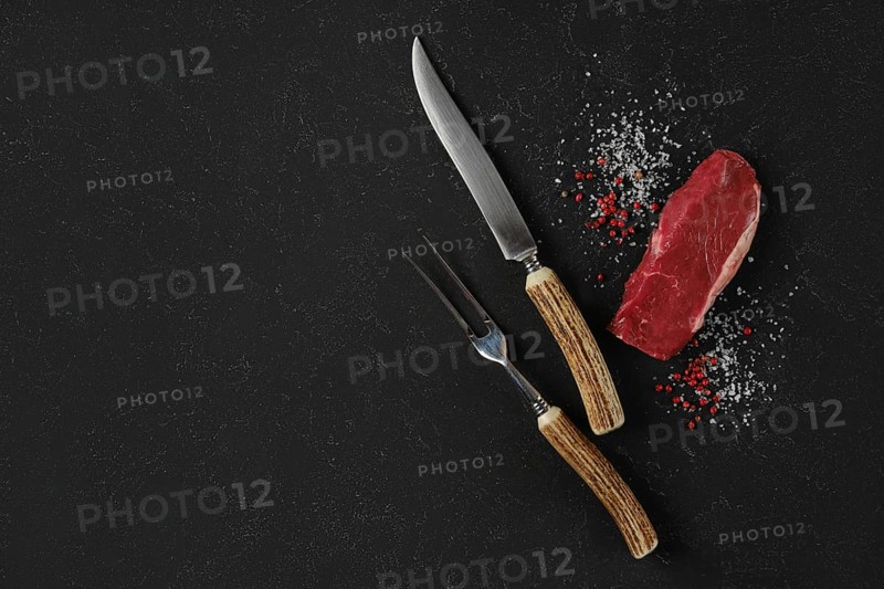 A knife and fork, with horn handles, rest beside a raw beef steak on a dark surface. Salt and pink peppercorn surround the meat, suggesting preparation for cooking or a gourmet meal