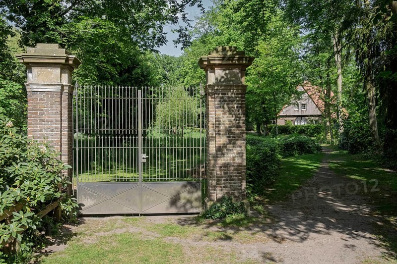 A wrought-iron gate between two brick pillars in a green, shady landscape with a building in the background, Welbergen, MÃ¼nsterland, North Rhine-Westphalia, Germany