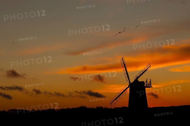 Windmill silhouette at sunset with a red sky and a skein or flock of Pink-footed geese (Anser brachyrhynchus) birds flying above, Burnham Ovary Staithe, Norfolk, England, United Kingdom