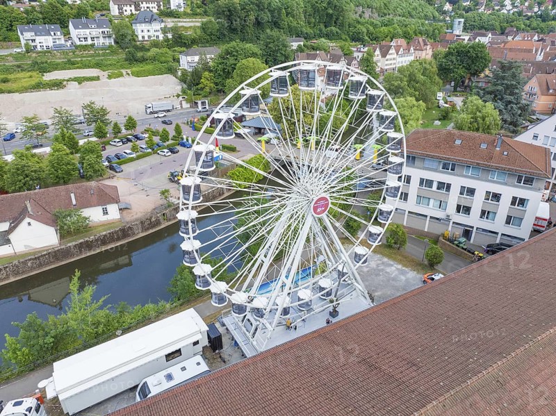 A white Ferris wheel next to a river, surrounded by buildings and green landscape, fire brigade exercise for gondola rescue, Germany