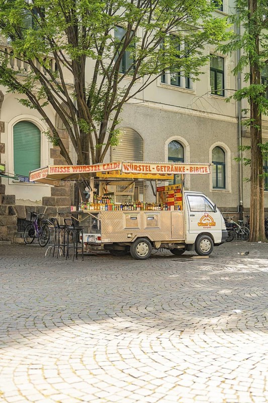 A food truck stands under trees on a paved square on a sunny day, Merano, South Tyrol, Italy