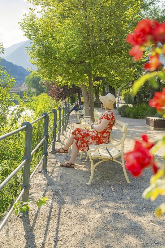 Woman in summer dress sitting on park bench, surrounded by flowers in relaxed atmosphere, Merano, South Tyrol, Italy