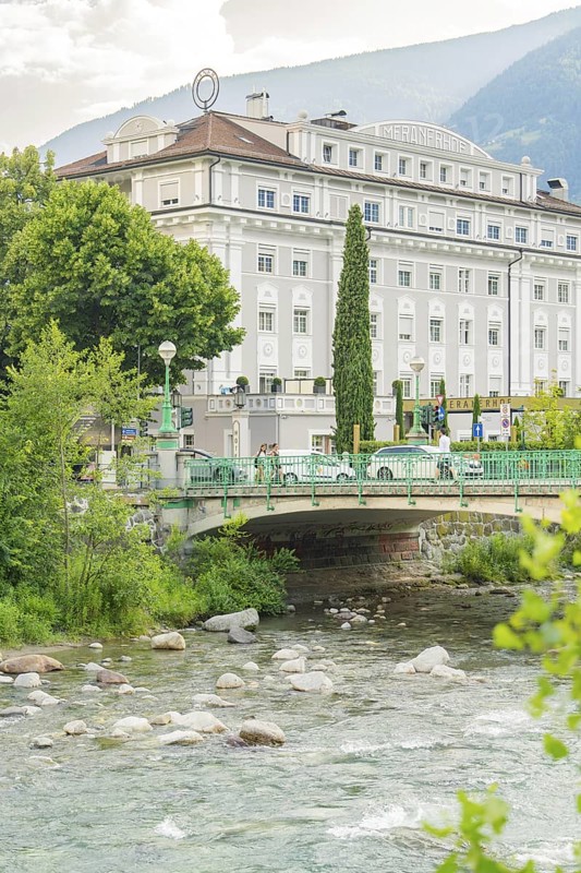 Historic riverside hotel with bridge and lush vegetation against a mountain backdrop, Merano, South Tyrol, Italy