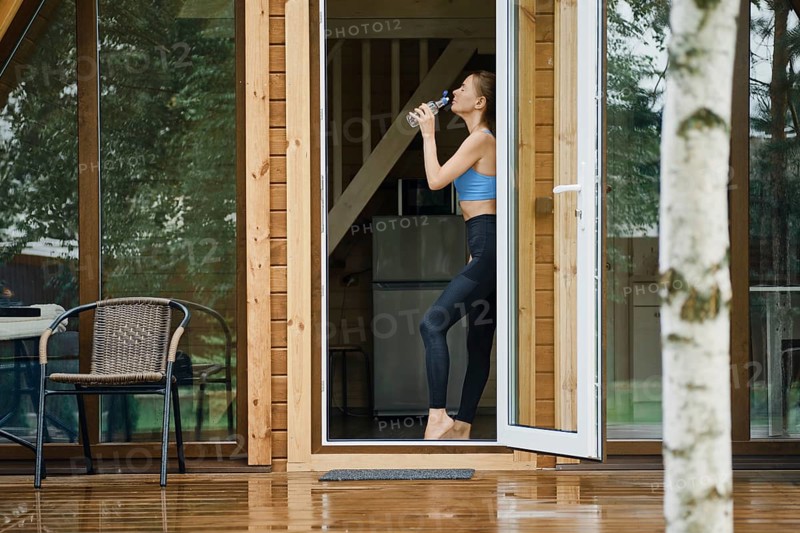 A fit woman in athletic wear stands in the doorway of her tiny wooden house, hydrating after a workout